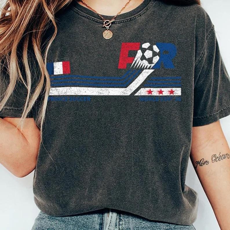 K&ouml;nnte beinhalten: Dunkelgraues T-Shirt mit einem Grafikdesign mit den Buchstaben "FR", einem Fu&szlig;ball und einem stilisierten Flaggen-Design in Rot, Wei&szlig; und Blau. Das T-Shirt hat einen Vintage-Look.