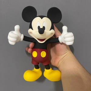 May include: A Mickey Mouse figurine featuring a black head with large round ears, a cream-colored face, and a black nose. The character wears a black shirt, dark red shorts with two yellow oval buttons, and large yellow shoes, with white gloved hands.
