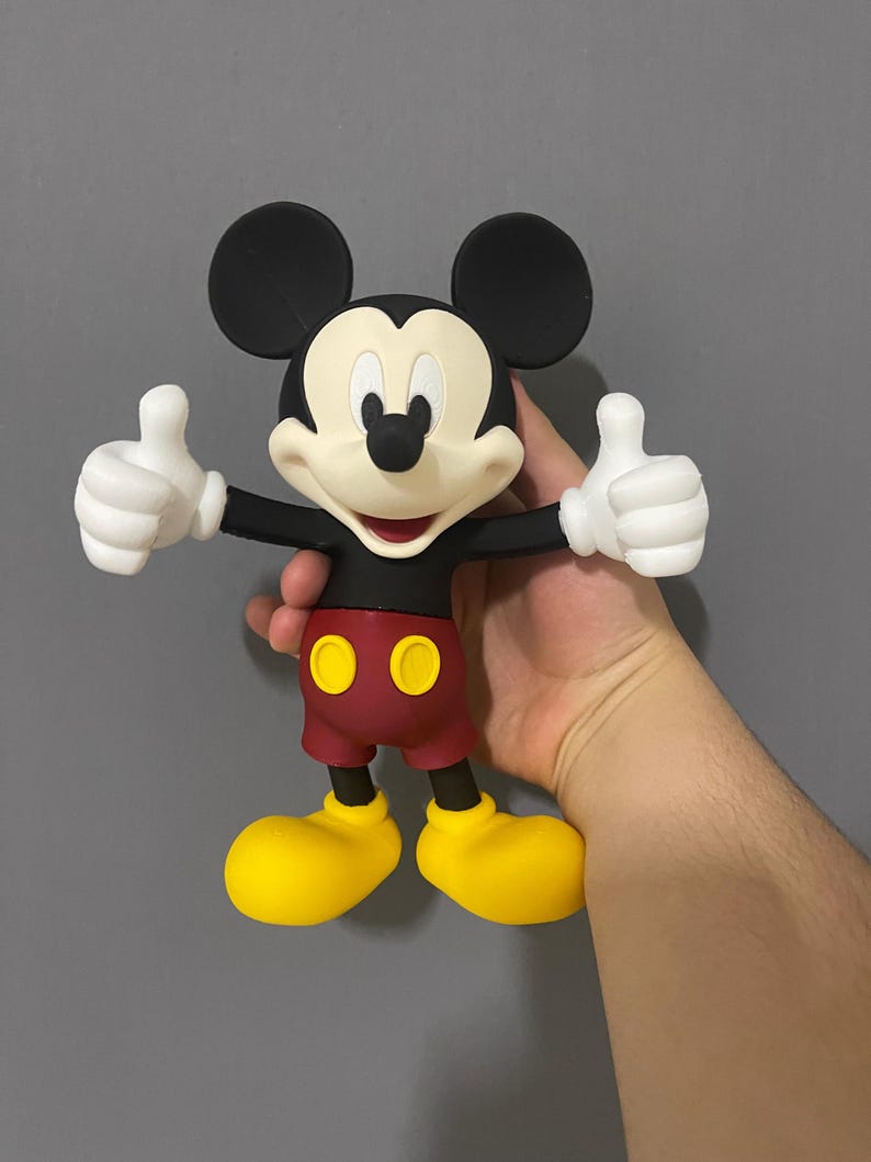 May include: A Mickey Mouse figurine featuring a black head with large round ears, a cream-colored face, and a black nose. The character wears a black shirt, dark red shorts with two yellow oval buttons, and large yellow shoes, with white gloved hands.