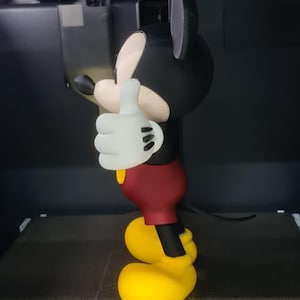 May include: A 3D printed figurine of a cartoon mouse character with black ears, a cream-colored face, white gloves, red shorts, and yellow shoes, standing in a thumbs-up pose. The figure is positioned inside a Bambu Lab 3D printer.