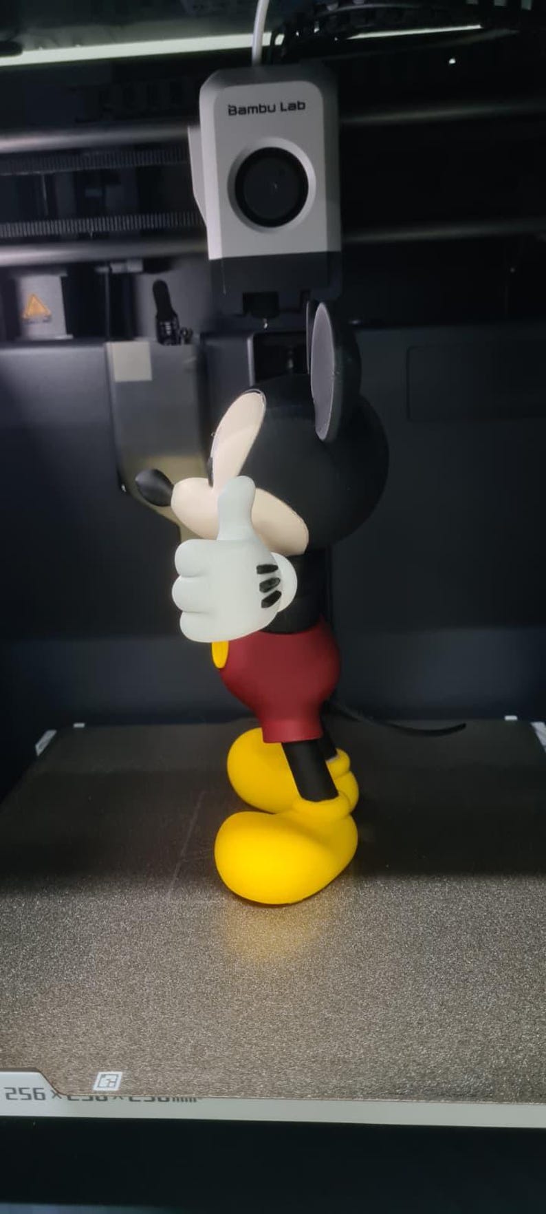 May include: A 3D printed figurine of a cartoon mouse character with black ears, a cream-colored face, white gloves, red shorts, and yellow shoes, standing in a thumbs-up pose. The figure is positioned inside a Bambu Lab 3D printer.