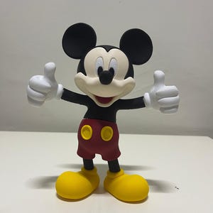 May include: A three-dimensional figurine of Mickey Mouse featuring a black head with large round ears, a cream-colored face, a black nose, and a wide smile. The character wears a black shirt, red shorts with two yellow buttons, and yellow shoes.