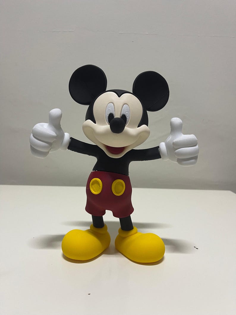 May include: A three-dimensional figurine of Mickey Mouse featuring a black head with large round ears, a cream-colored face, a black nose, and a wide smile. The character wears a black shirt, red shorts with two yellow buttons, and yellow shoes.