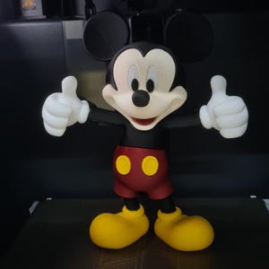 May include: A 3D printed Mickey Mouse figurine with black ears, a cream face, red shorts with two yellow buttons, and yellow shoes, standing inside a Bambu Lab printer with a camera unit above and a build volume label reading 256.
