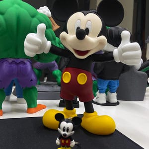 May include: A large Mickey Mouse figurine with black ears, a cream face, red shorts with two yellow buttons, and yellow shoes, standing with both thumbs up. In front of it sits a smaller Mickey Mouse keychain with the same color scheme.