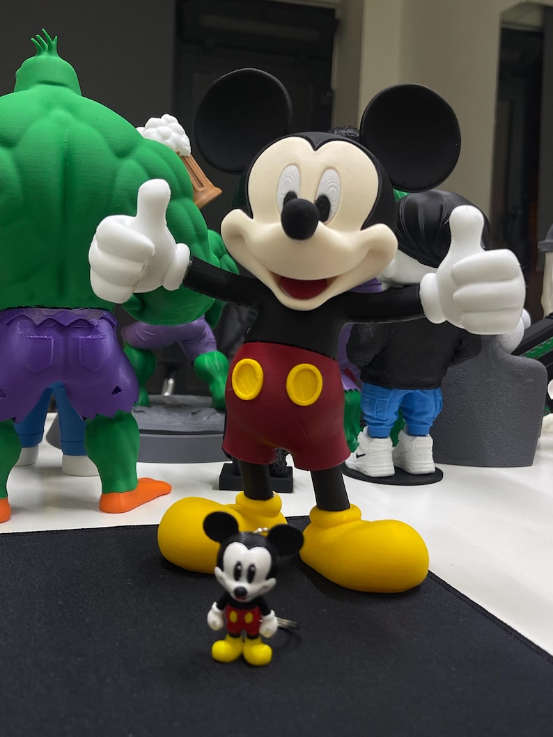May include: A large Mickey Mouse figurine with black ears, a cream face, red shorts with two yellow buttons, and yellow shoes, standing with both thumbs up. In front of it sits a smaller Mickey Mouse keychain with the same color scheme.