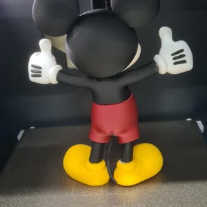 May include: A 3D printed figurine of a cartoon mouse with large black ears, a black head and torso, red shorts, and yellow shoes. The figure has white gloves with three black lines on each, and both arms are raised with thumbs pointing upward.