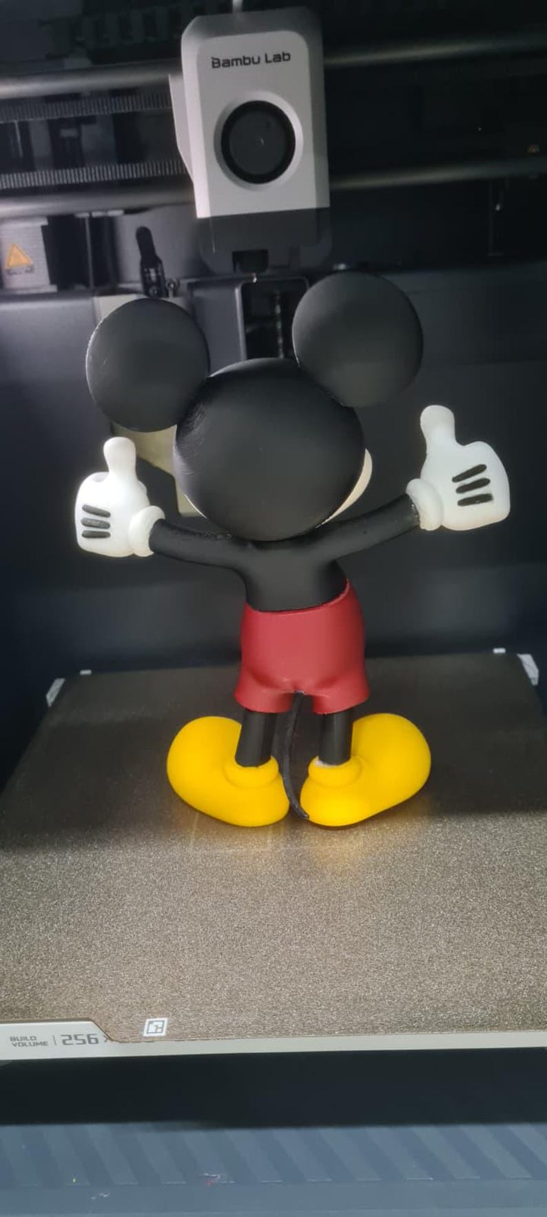 May include: A 3D printed figurine of a cartoon mouse with large black ears, a black head and torso, red shorts, and yellow shoes. The figure has white gloves with three black lines on each, and both arms are raised with thumbs pointing upward.