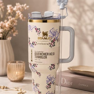 Pu&ograve; includere: Bicchiere Stanley Quencher H2.0 color crema con un motivo floreale e fiocchi. Presenta un manico grigio, una cannuccia trasparente e un accessorio per cannuccia a forma di fiocco. Il bicchiere contiene 1,18 L e riporta la scritta "LOVESHACK" e "QUENCHER H2.0 TUMBLER".