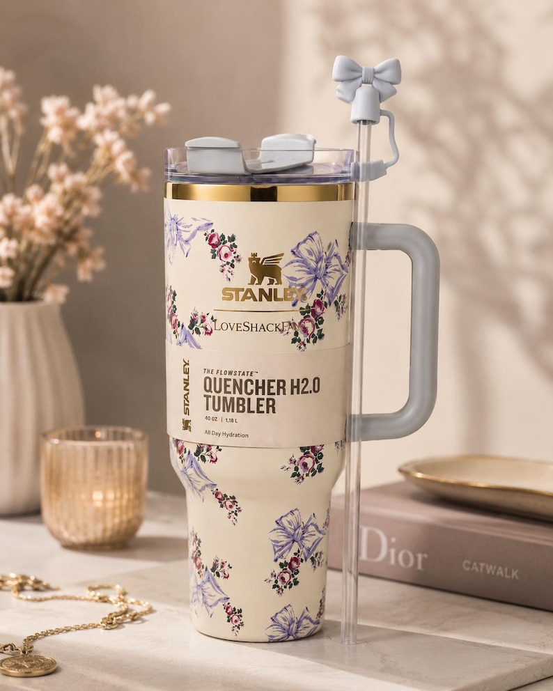 Pu&ograve; includere: Bicchiere Stanley Quencher H2.0 color crema con un motivo floreale e fiocchi. Presenta un manico grigio, una cannuccia trasparente e un accessorio per cannuccia a forma di fiocco. Il bicchiere contiene 1,18 L e riporta la scritta "LOVESHACK" e "QUENCHER H2.0 TUMBLER".