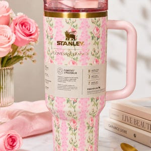 May include: A floral-patterned Stanley tumbler with a pink handle and lid. The tumbler features a cream background with pink roses and green leaves. The text "STANLEY LOVESHACKFANCY" is visible on the tumbler. The tumbler is designed to keep drinks cold for 2 days.