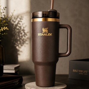 Puede incluir: Un vaso Stanley marr&oacute;n con asa y pajita. El vaso tiene un borde dorado y el logotipo de Stanley en dorado. Se encuentra sobre un posavasos de m&aacute;rmol. Dise&ntilde;ado para mantener las bebidas fr&iacute;as o calientes durante horas.