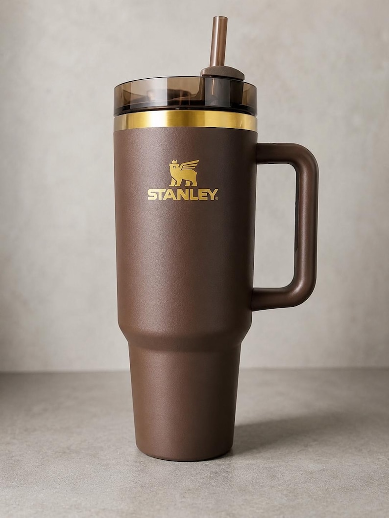 Puede incluir: Un termo de viaje Stanley marr&oacute;n con asa, pajita y borde dorado. El logotipo de Stanley est&aacute; impreso en dorado en la parte delantera del termo. El termo est&aacute; dise&ntilde;ado para mantener las bebidas calientes o fr&iacute;as.