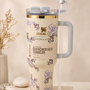 Pu&ograve; includere: Bicchiere Stanley Quencher H2.0 color crema con motivo floreale e fiocchi. Presenta un manico grigio, una cannuccia trasparente con un fiocco e il testo "QUENCHER H2.0 TUMBLER". Il bicchiere contiene 1,18L.