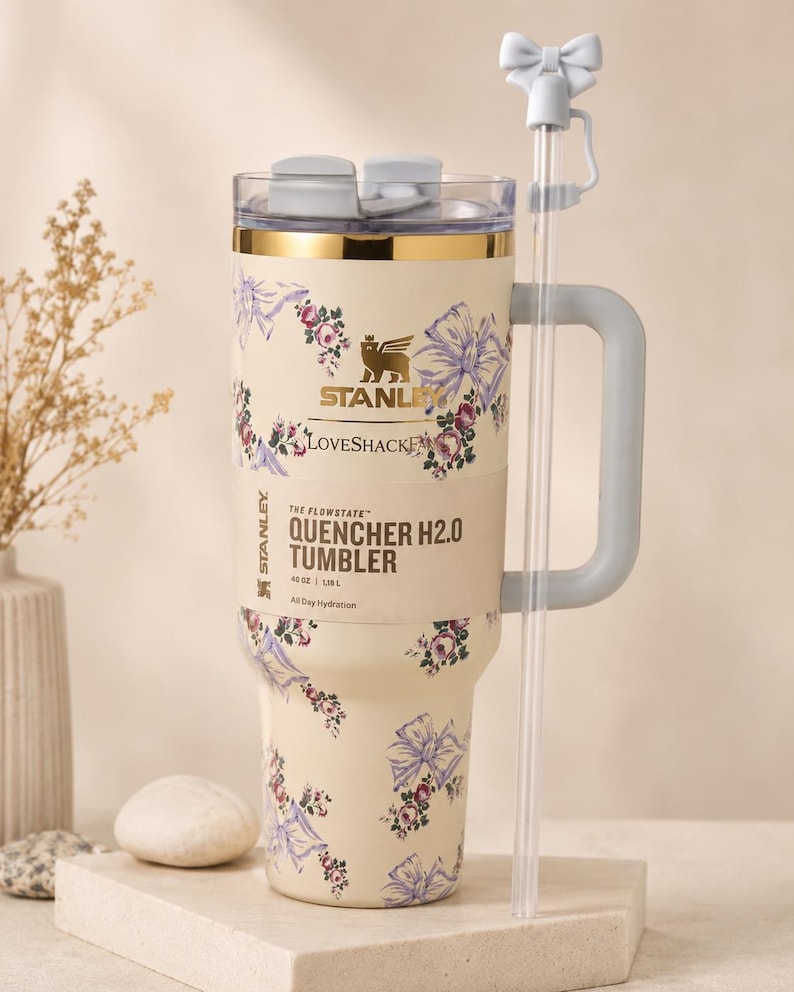 Pu&ograve; includere: Bicchiere Stanley Quencher H2.0 color crema con motivo floreale e fiocchi. Presenta un manico grigio, una cannuccia trasparente con un fiocco e il testo "QUENCHER H2.0 TUMBLER". Il bicchiere contiene 1,18L.