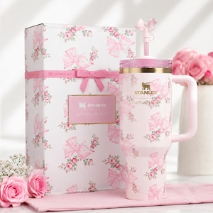 May include: A pink Stanley tumbler and gift box set with a floral and bow pattern. The tumbler has a handle and straw, and the box is tied with a pink ribbon. The set is decorated with roses and the text "Stanley Loveshackfancy".