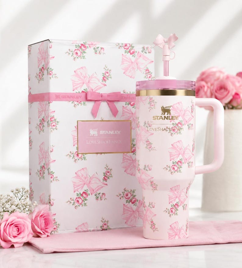 May include: A pink Stanley tumbler and gift box set with a floral and bow pattern. The tumbler has a handle and straw, and the box is tied with a pink ribbon. The set is decorated with roses and the text "Stanley Loveshackfancy".