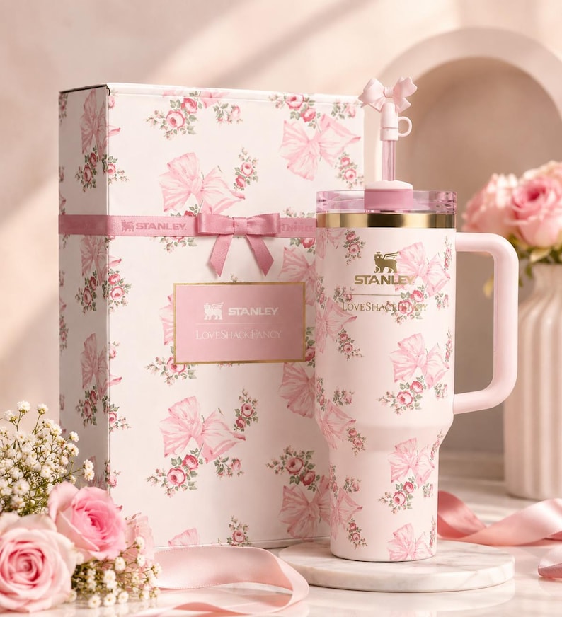 May include: A pink Stanley Loveshack Fancy tumbler and gift box set. The tumbler and box feature a floral and bow pattern. The tumbler has a handle and a straw. The gift box is tied with a pink ribbon.