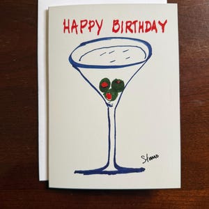 Puede incluir: Tarjeta de cumpleaños con un dibujo a mano de una copa de martini llena de aceitunas verdes y las palabras "HAPPY BIRTHAY" en rojo. La copa de martini está delineada en azul, sobre fondo blanco.