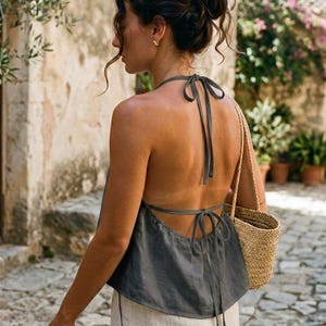 Könnte beinhalten: Ein graues Neckholder-Top mit Bindebändern am Hals und Rücken. Das Top wird mit hellen Hosen und einer kleinen gewebten Tasche kombiniert. Das Bild ist im Freien aufgenommen, mit einer Steinmauer und Grün in der Hintergrund.