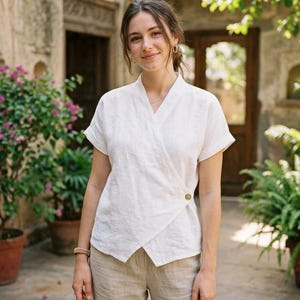 May include: A white linen wrap top with short sleeves and a single button closure. The top is paired with beige linen pants. The outfit is worn by a person in an outdoor setting with plants and a stone building in the background.