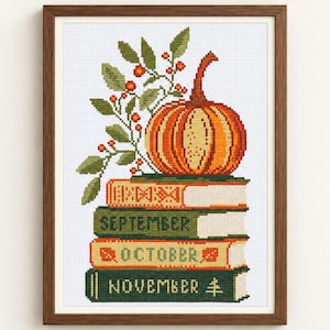 May include: A framed cross-stitch embroidery featuring a stack of three books labeled September, October, and November. An orange pumpkin sits atop the books, accompanied by a sprig of green leaves and small red berries, all set against a white grid.