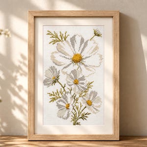 May include: A framed cross-stitch embroidery featuring a botanical illustration of white cosmos flowers with yellow centres and green foliage on a white fabric background, enclosed in a light-toned wooden frame with a clean, rectangular design.
