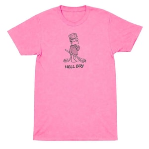 May include: A bright pink t-shirt featuring a black and white cartoon illustration of a character holding a pitchfork. The text "HELL BOY" is printed below the illustration. The shirt is a solid color.