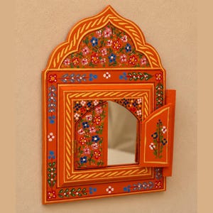 May include: An orange wall mirror with a pointed arch top and two small hinged doors. The frame is decorated with hand-painted floral patterns in red, blue, white, and green. The central mirror is rectangular with an arched top section.