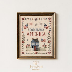 May include: Framed cross-stitch artwork featuring a blue house with a red door and American flags. The text "GOD BLESS AMERICA" is displayed above the house. The artwork is surrounded by floral and star motifs, and is in a brown wooden frame.