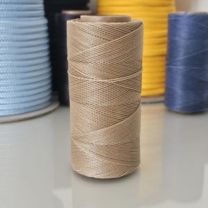 Puede incluir: Un carrete de hilo beige es el punto focal, con otros carretes de hilo en varios colores, incluyendo azul, amarillo y turquesa, en el fondo. El hilo está bien enrollado, y los carretes están dispuestos sobre una superficie reflectante. Esta imagen muestra una variedad de hilos.