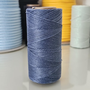 Puede incluir: Un carrete de cordón encerado azul grisáceo. Otros carretes de cordón en varios colores, incluyendo azul claro, amarillo, blanco y verde azulado, son visibles en el fondo. El cordón es para manualidades.