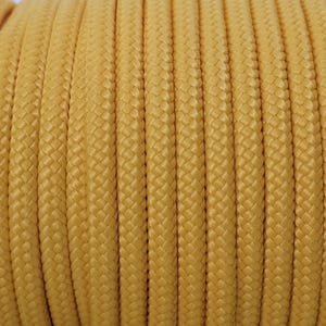 Peut inclure: Gros plan d'une bobine de corde tressée jaune. La corde est enroulée serrée, montrant une texture détaillée de brins entrelacés. La couleur est un jaune doré chaud, et la corde semble durable.