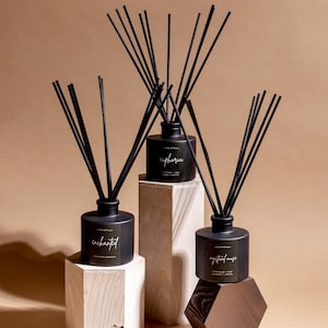 May include: Three black reed diffusers with black reeds are displayed on wooden blocks and a geometric stand. Each diffuser has a label with a different name: "enchanted", "euphoria", and "mystical muse". A pine cone and a small piece of wood are in the foreground.