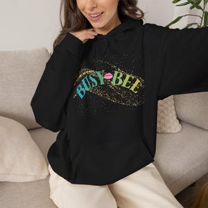 Op de afbeelding: Een zwarte hoodie met een grafisch ontwerp met de tekst BUSY BEE in turquoise en gele letters. Het ontwerp bevat een roze lippenicoon tussen de woorden en een gouden glittereffect-patroon over het borstgedeelte van het kledingstuk.