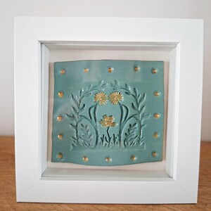 May include: A square white frame containing a teal ceramic tile with embossed floral designs. The tile features two dandelion heads and a small flower in gold, surrounded by leafy stems. Small gold dots line the perimeter of the tile.