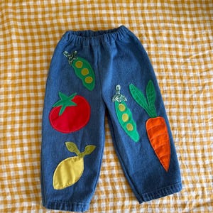 May include: Blue denim pants with colorful fabric appliqués of vegetables. The appliqués include a red tomato, a yellow lemon, green pea pods, and an orange carrot. The pants have an elastic waistband.