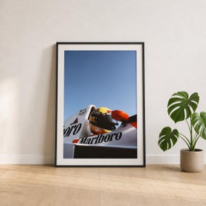 May include: Framed print featuring a Formula 1 race car with a yellow and black helmet. The car is white with red and black accents and the "Marlboro" logo. The background is a clear blue sky.