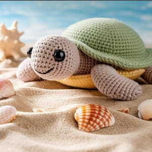 May include: A crocheted turtle toy with a green shell, gray body, and black eyes rests on sand, surrounded by seashells. The turtle has a smiling face and is a handmade item.