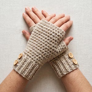 Op de afbeelding: Beige gehaakte vingerloze handschoenen met geribbelde manchetten en houten knopen. De handschoenen zijn handgemaakt en hebben een gestructureerd steekpatroon. De knopen zijn rond en voegen een decoratief element toe. De handschoenen houden de handen warm.
