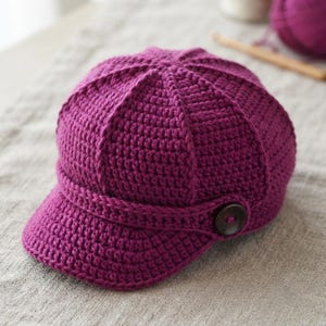 May include: A deep magenta crocheted newsboy cap featuring a structured crown with vertical ridges, a short stiffened brim, and a decorative horizontal band across the front secured by a single dark brown circular button on the right side.