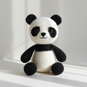 Könnte beinhalten: Ein handgehäkelter Panda-Bär mit weißem Körper und schwarzen Akzenten. Der Panda hat schwarze Ohren, Augen, Arme und Füße. Das Gesicht hat eine schwarze Nase und ein kleines, gesticktes Lächeln. Das Spielzeug sitzt aufrecht.