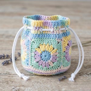 May include: A small drawstring pouch crafted from crocheted granny squares in pastel shades of yellow, light blue, mint green, and lavender. The bag features a white cord closure threaded through the top edge, creating a gathered opening.