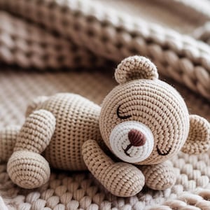 Könnte beinhalten: Ein gehäkelter Teddybär in Hellbraun, der auf einer passenden Strickdecke liegt. Der Bär hat eine weiße Schnauze, eine braune Nase und geschlossene Augen. Die Decke hat ein strukturiertes, gewebtes Aussehen.