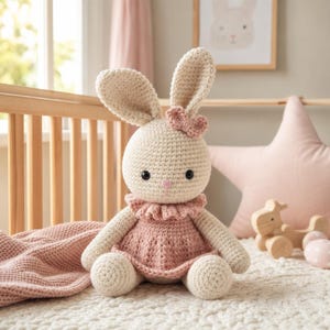May include: A crocheted bunny doll in cream and pink. The bunny has long ears, a pink bow, and a pink dress with a ruffled collar. The doll is sitting on a white surface with a pink blanket nearby. A wooden crib is in the background.