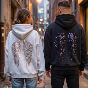 Pode incluir: Duas camisolas com capuz, uma cinza claro e outra preta, cada uma com asas de anjo com lantejoulas nas costas. A camisola cinza claro tem asas prateadas, a preta tem asas roxas. Ambas têm cordões.