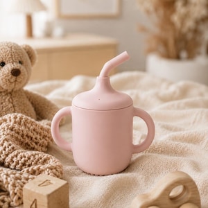Pode incluir: Uma chávena rosa claro com uma palhinha curva e duas pegas. A chávena é feita de um material mate e está sobre um cobertor creme, ao lado de um carro de brinquedo de madeira e um urso de peluche.