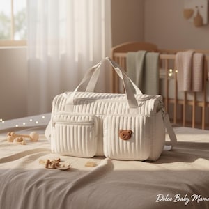 May include: Cream-colored quilted baby bag with a teddy bear patch on the front pocket. The bag has two exterior pockets, a zipper closure, and a shoulder strap. The bag is sitting on a bed with wooden toys nearby.