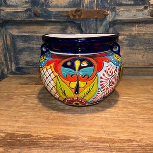 May include: A round, hand-painted ceramic pot with a vibrant floral design. The pot features a dark blue rim and handles, with a colorful pattern of red, yellow, blue, and green. The pot is sitting on a wooden surface.
