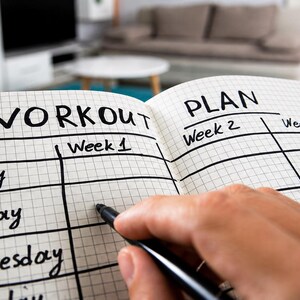 May include: A workout plan notebook open, showing sections for each day of the week and weeks 1, 2, and 3. The word "WORKOUT" is written at the top, with a hand holding a pen, ready to fill in the plan.